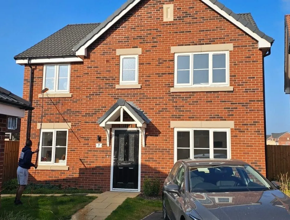 An operative cleaning the first-floor windows of a new-build house from the front garden